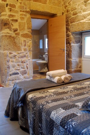Una habitación con paredes de piedra, una cama, una puerta de madera y vistas a otra habitación con bañera y retrete.