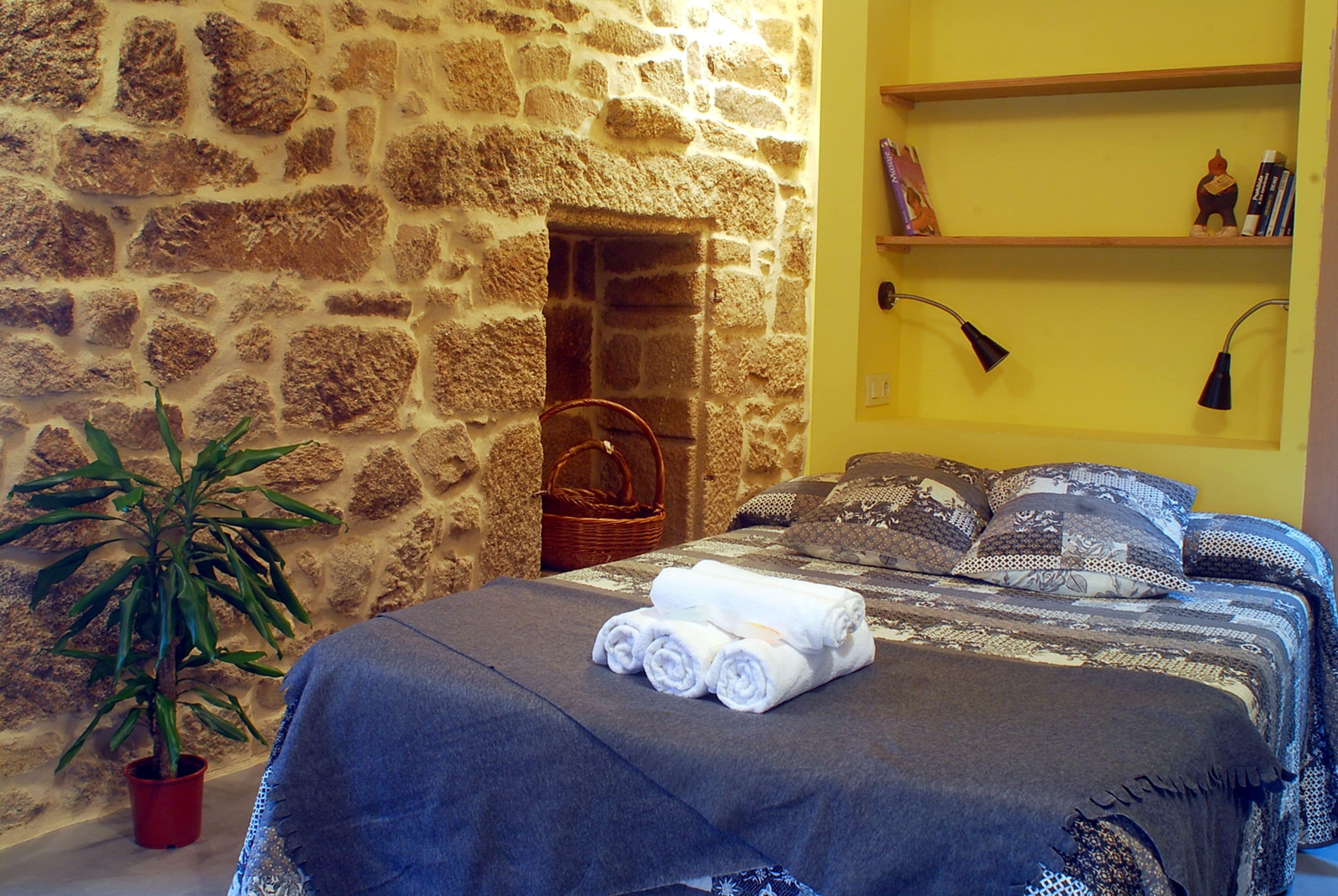 A bedroom with a stone wall, a bed with rolled towels, a potted plant, and a shelf with books and a lamp.