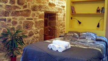 A bedroom with a stone wall, a bed with rolled towels, a potted plant, and a shelf with books and a lamp.