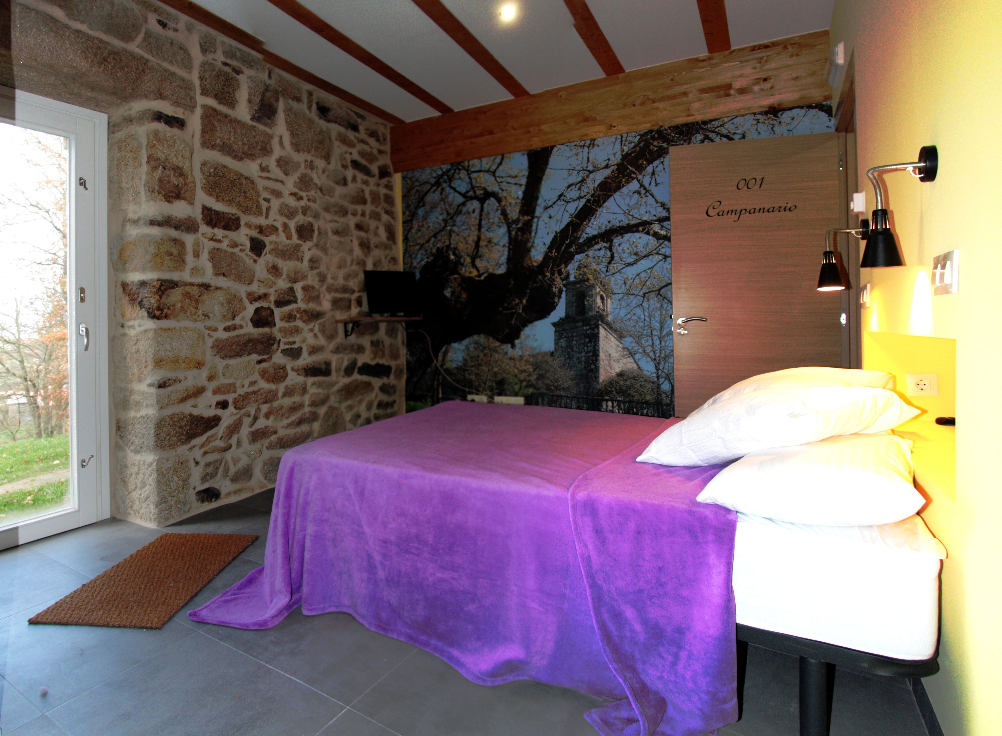 A bedroom with a stone wall, a bed with a purple bedspread, a white pillow, and a nightstand with a lamp.