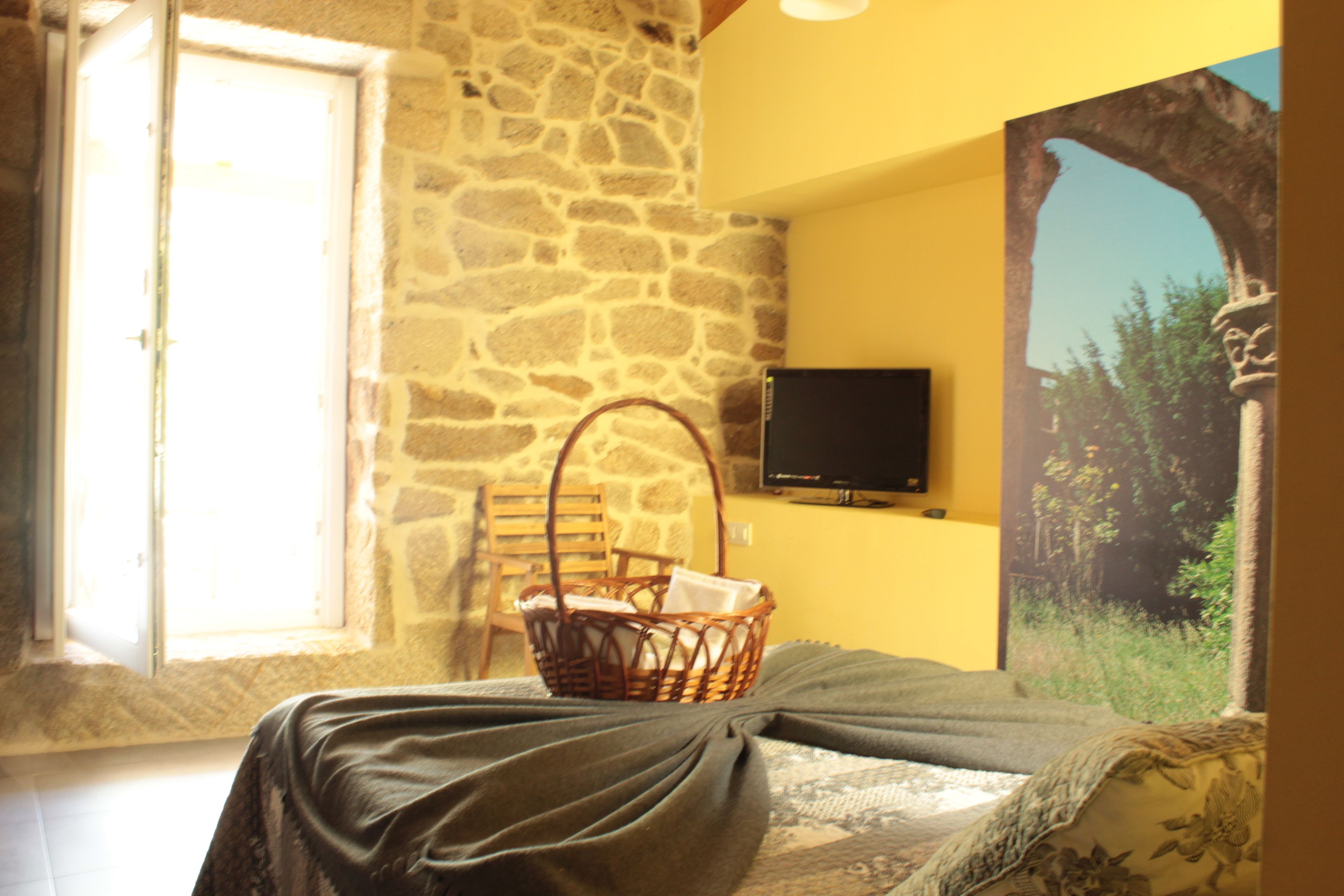 A room with a stone wall, a bed with a dark bedspread, a wicker basket, a television on the wall, and a window with natural light.