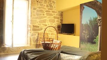 A room with a stone wall, a bed with a dark bedspread, a wicker basket, a television on the wall, and a window with natural light.