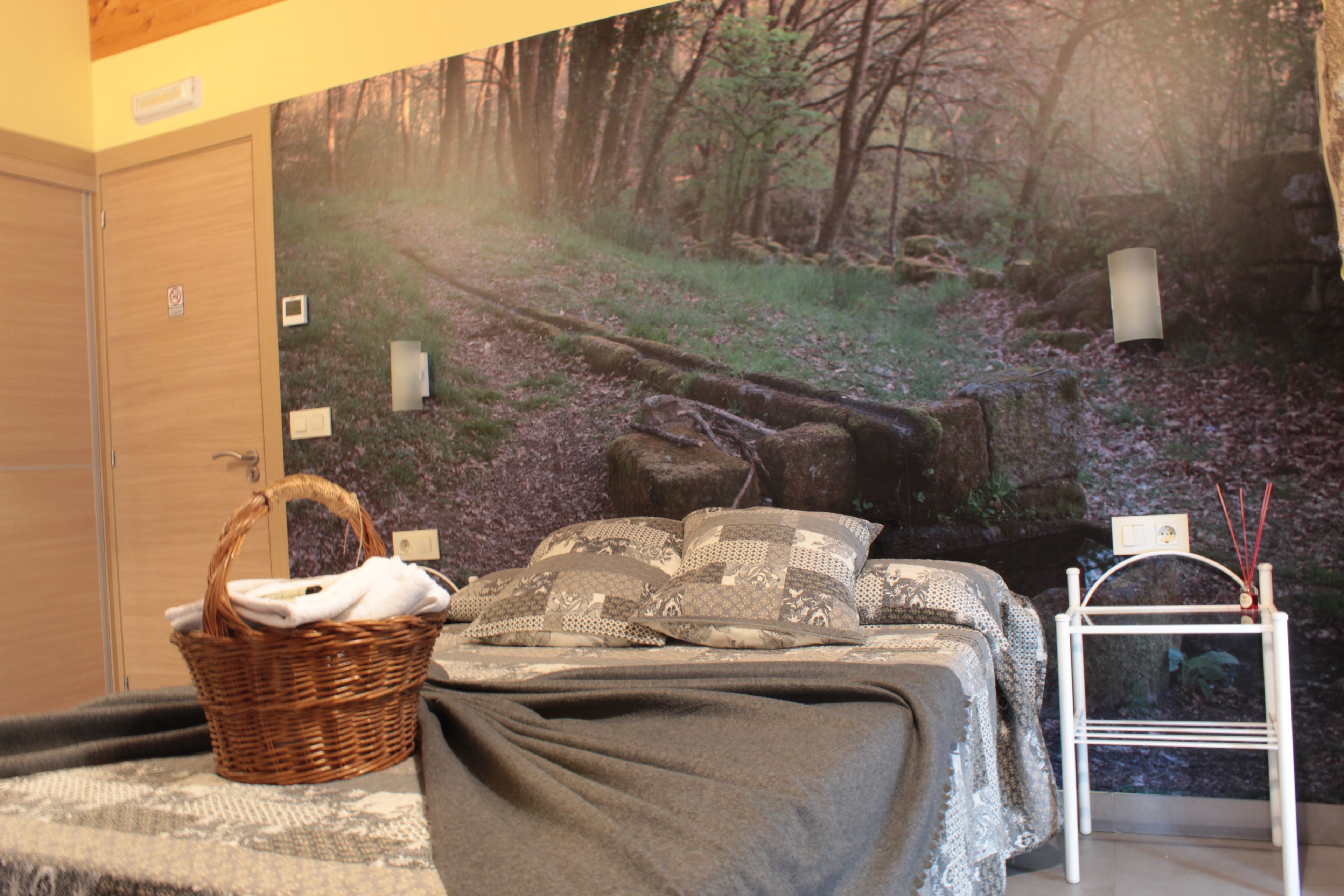 A bedroom with a bed, a wicker basket, a white nightstand, and a forest-themed wall mural.