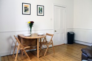 A dining area with a wooden table, two chairs, a vase with flowers, and two framed pictures on the wall.