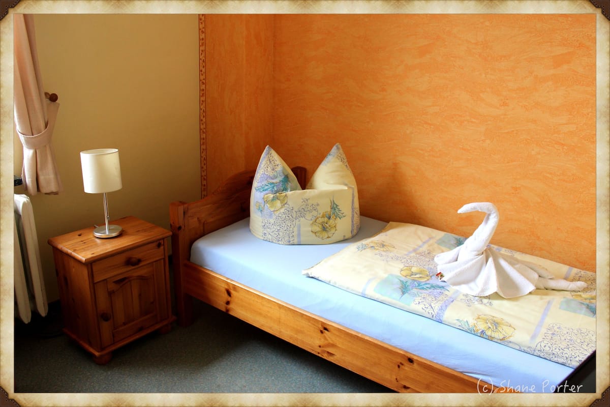 A single bed with a blue mattress, two pillows with floral designs, a wooden nightstand with a lamp, and a towel shaped like a swan.