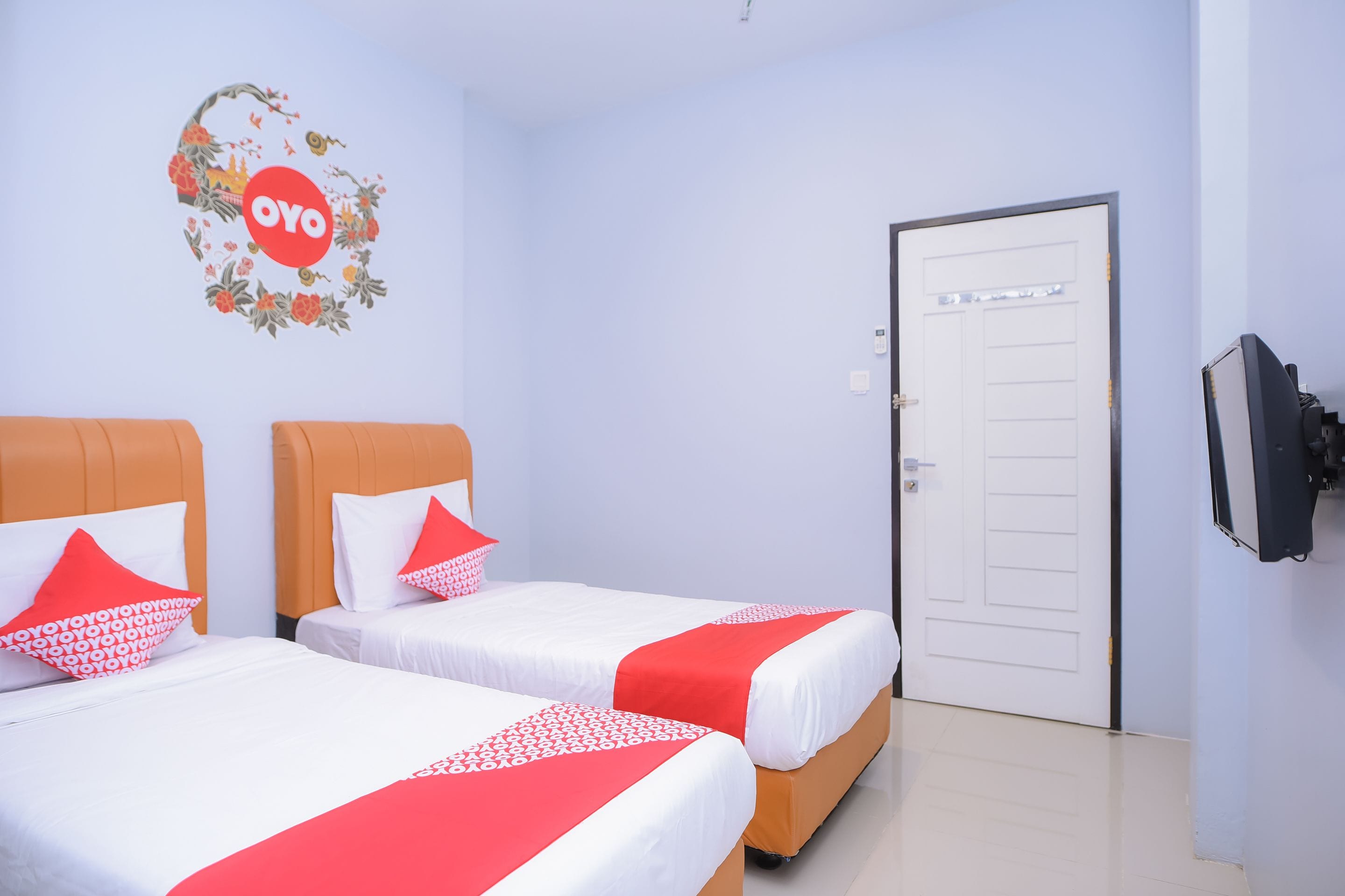 A hotel room with two beds, each with white bedding and red accents, a floral wall decal, and a television on the wall.