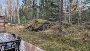A wooden deck with chairs, surrounded by a forest with tall trees and a moss-covered rock.