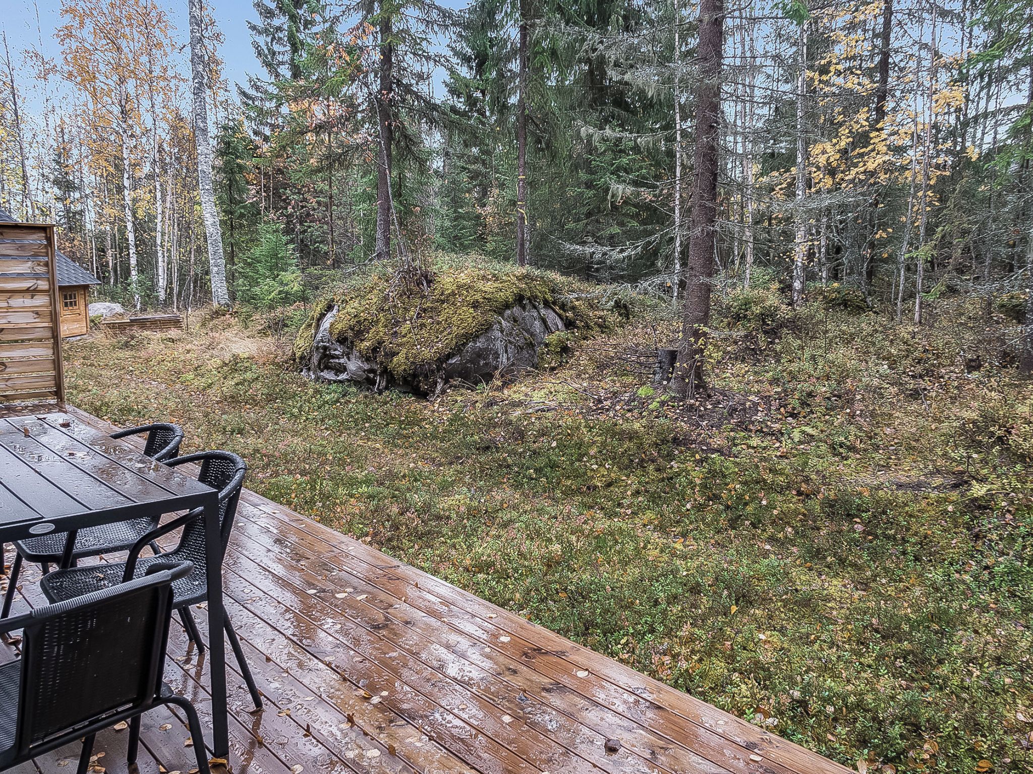 A wooden deck with a table and chairs, surrounded by a forested area with rocks and trees.