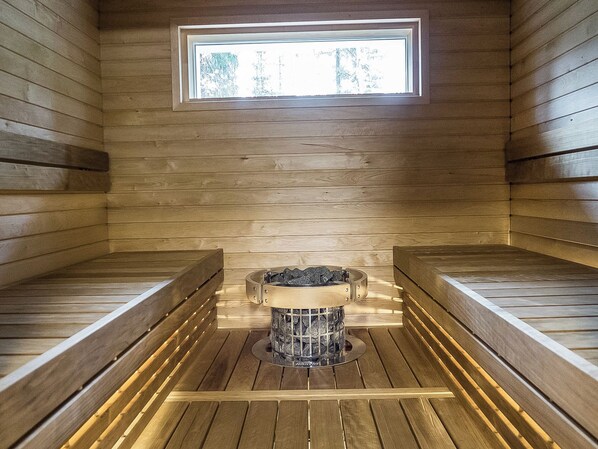 A sauna room with wooden benches, a central heating unit, and a window.