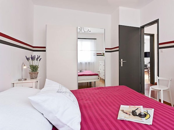 A bedroom with a bed, a bedside table with a lamp and a plant, a mirror, and a door leading to a bathroom.