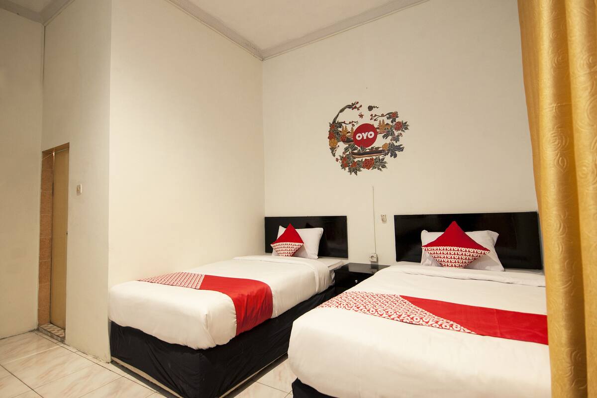 Une chambre d’hôtel avec deux lits, chacun doté d’une tête de lit noire et de draps blancs. Chaque lit est agrémenté d’un couvre-lit rouge et blanc à motifs. Un élément décoratif mural portant le mot « OYO » est visible au-dessus des lits.
