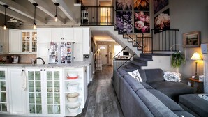 Un salón espacioso con un sofá gris, una cocina con armarios blancos y una escalera que sube a un nivel superior.