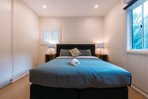 Un dormitorio con una cama grande, dos mesitas de noche, una ventana con persianas y vistas al exterior con vegetación.