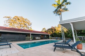 A swimming pool with lounge chairs, a covered patio, and a building with a red-tiled roof.