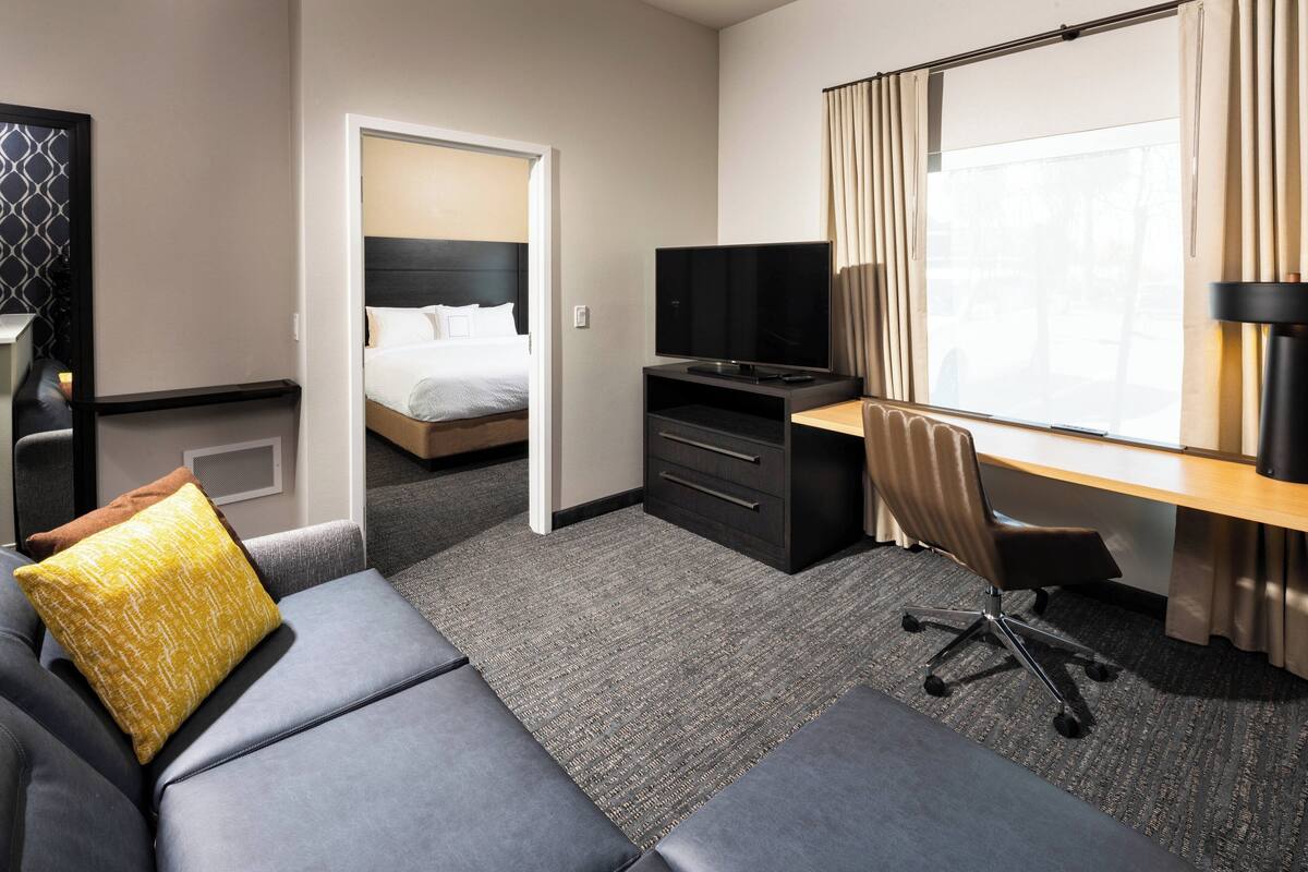 Ein Hotelzimmer mit Bett, Schreibtisch mit Fernseher, Sofa und großem Fenster mit Vorhängen.