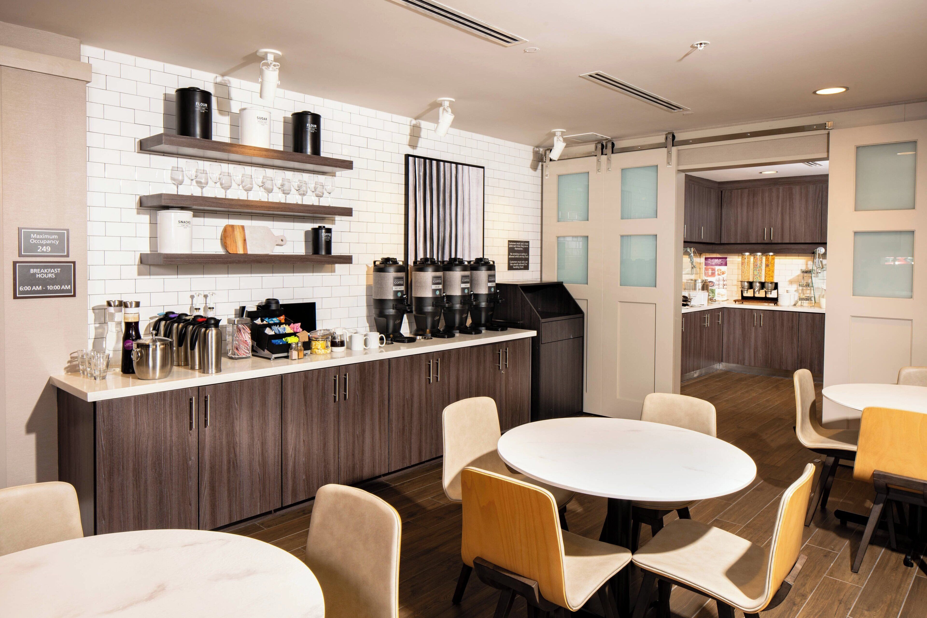Una cocina moderna con salpicadero de azulejos blancos, armarios de madera y una zona de café. Hay mesas redondas y sillas dispuestas para comer.