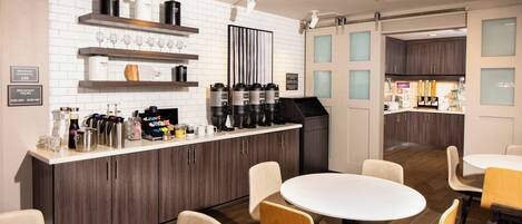 Una cocina moderna con salpicadero de azulejos blancos, armarios de madera y una zona de café. Hay mesas redondas y sillas dispuestas para comer.