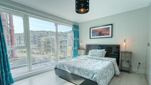 A modern bedroom with a large window, a bed with a grey and white patterned bedspread, a bedside table with a lamp, and a framed picture on the wall. - The Larry Mullen Suite - hiphipstay (Dublin)