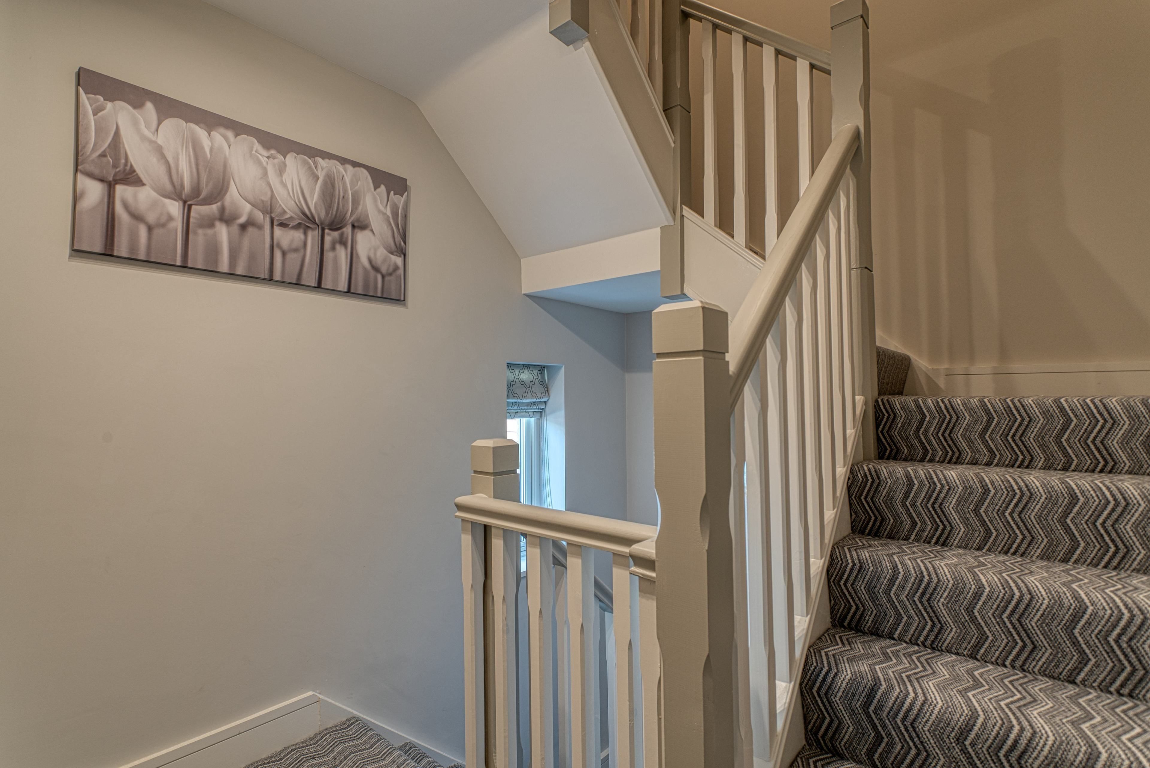 Un escalier recouvert d’une moquette à motifs, une rampe blanche et un tableau représentant des fleurs accroché au mur.