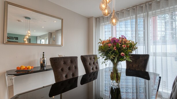 A dining area with a glass table, two tufted chairs, a vase with flowers, a mirror, and a window with curtains. - The Larry Mullen Suite - hiphipstay (Dublin)