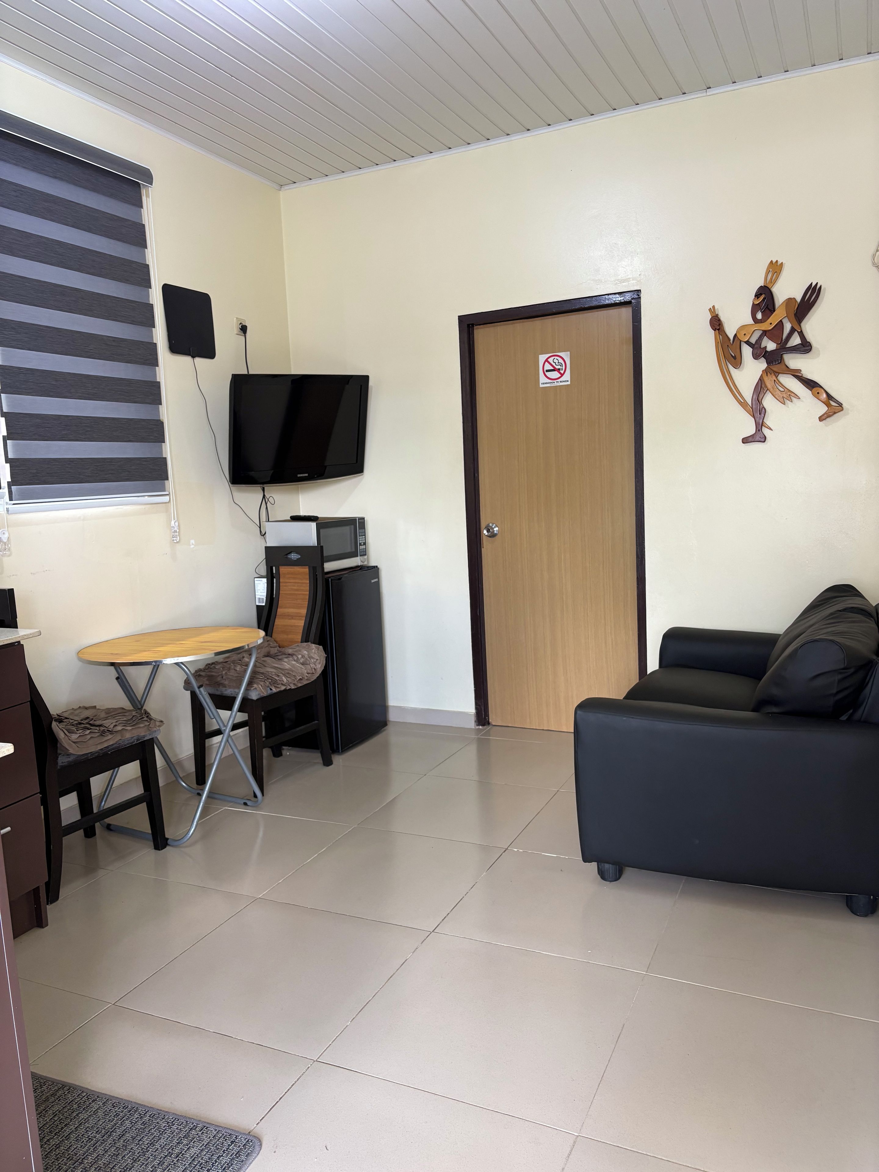 Une pièce comprenant une télévision, une table avec des chaises, un canapé, une porte avec un panneau « interdiction de fumer » et un élément de décoration fixé au mur.
