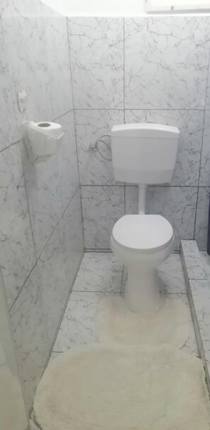 A bathroom with a toilet, a toilet paper holder, and a white rug on the floor. - DoksaTas Apartements A (Paramaribo)