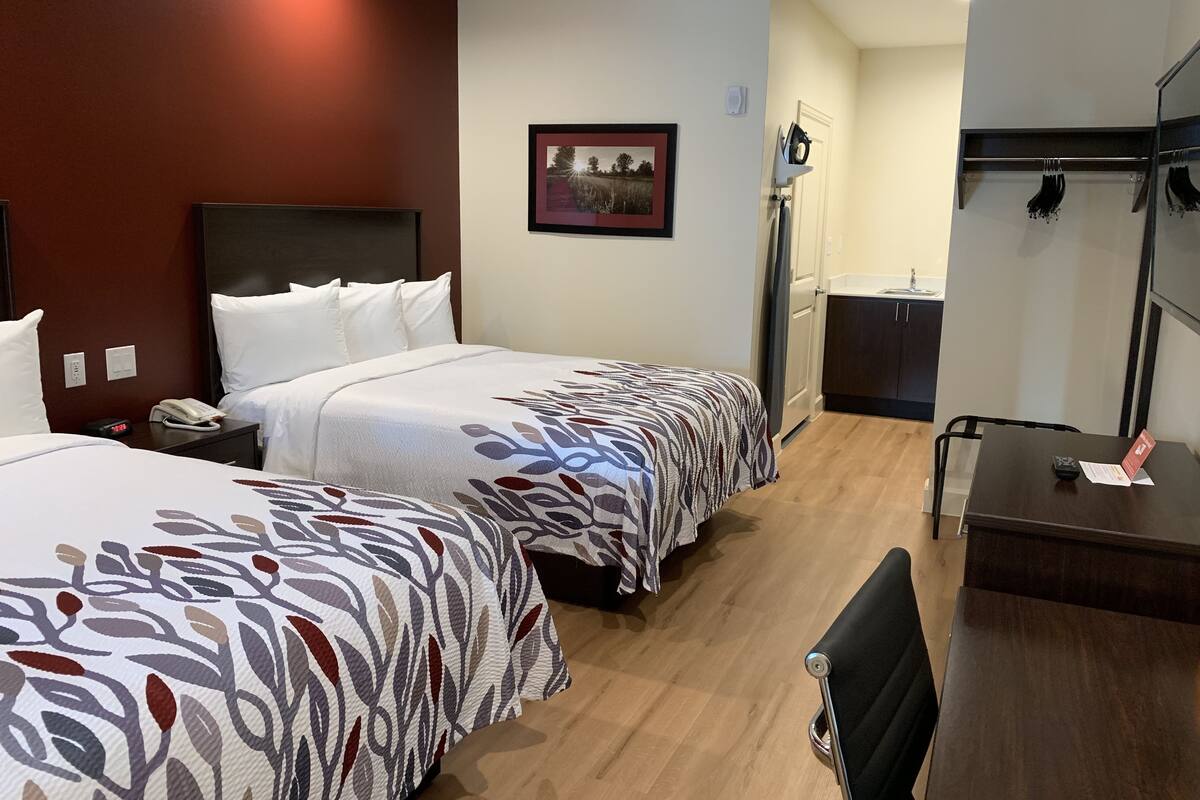 A hotel room with two beds, a desk, a chair, a mirror, a picture on the wall, and a small kitchenette.
