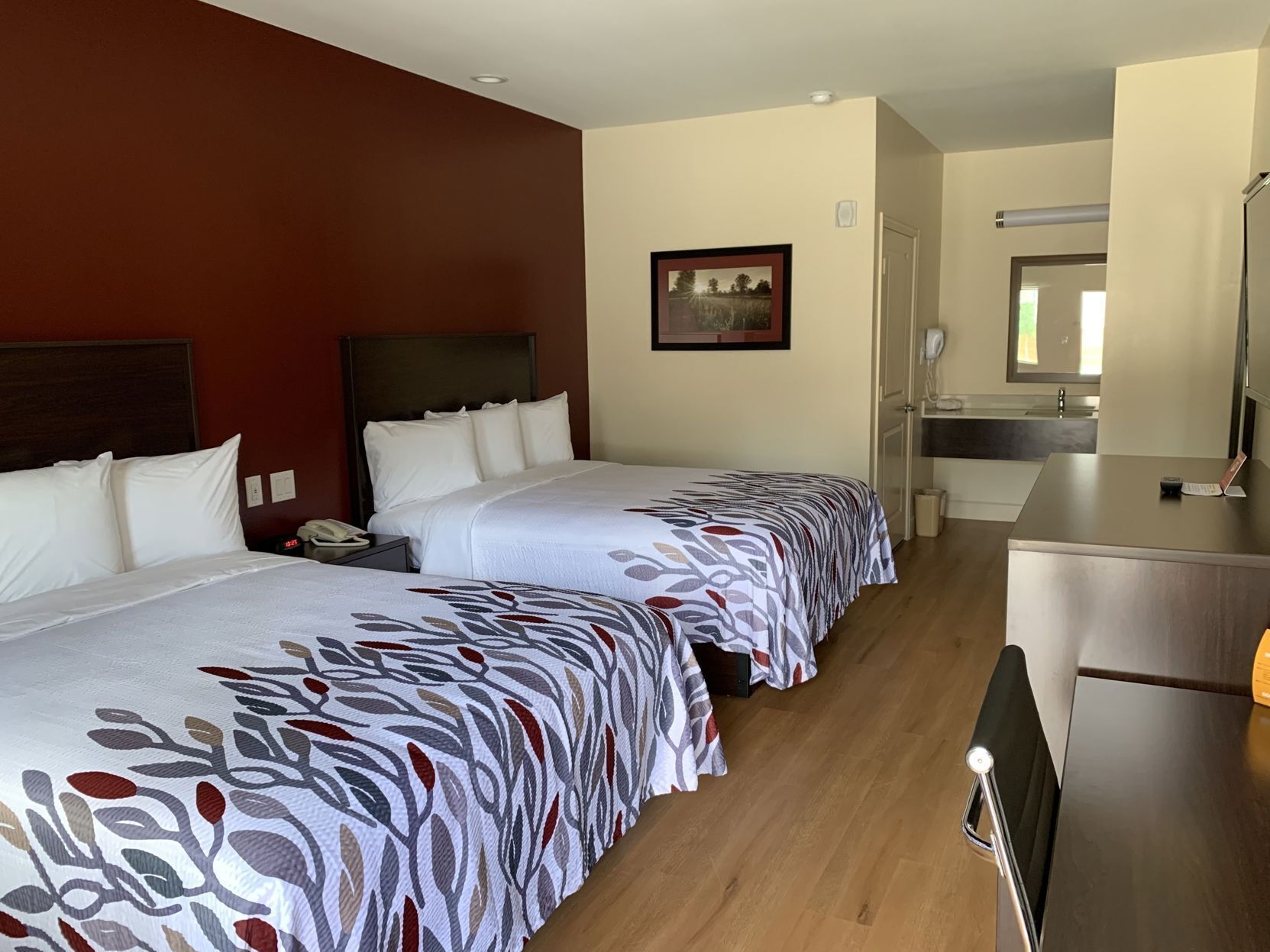 A hotel room with two beds, a desk, a chair, a bathroom, and a painting on the wall.