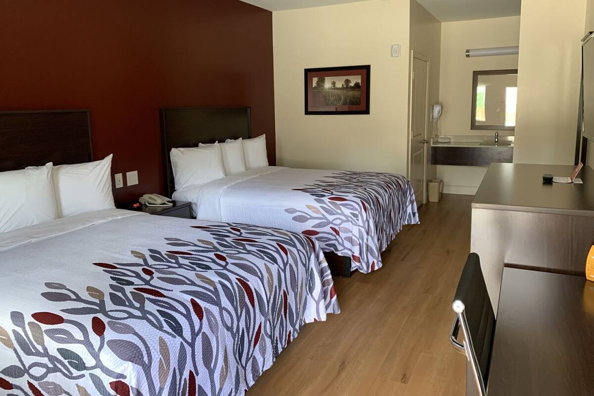 A hotel room with two beds, a desk, a chair, a bathroom, and a painting on the wall.