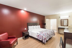 A hotel room with a bed, a nightstand, a red chair, a desk with a mirror, and a door. - Red Roof Inn Houston - Willowbrook (Houston)