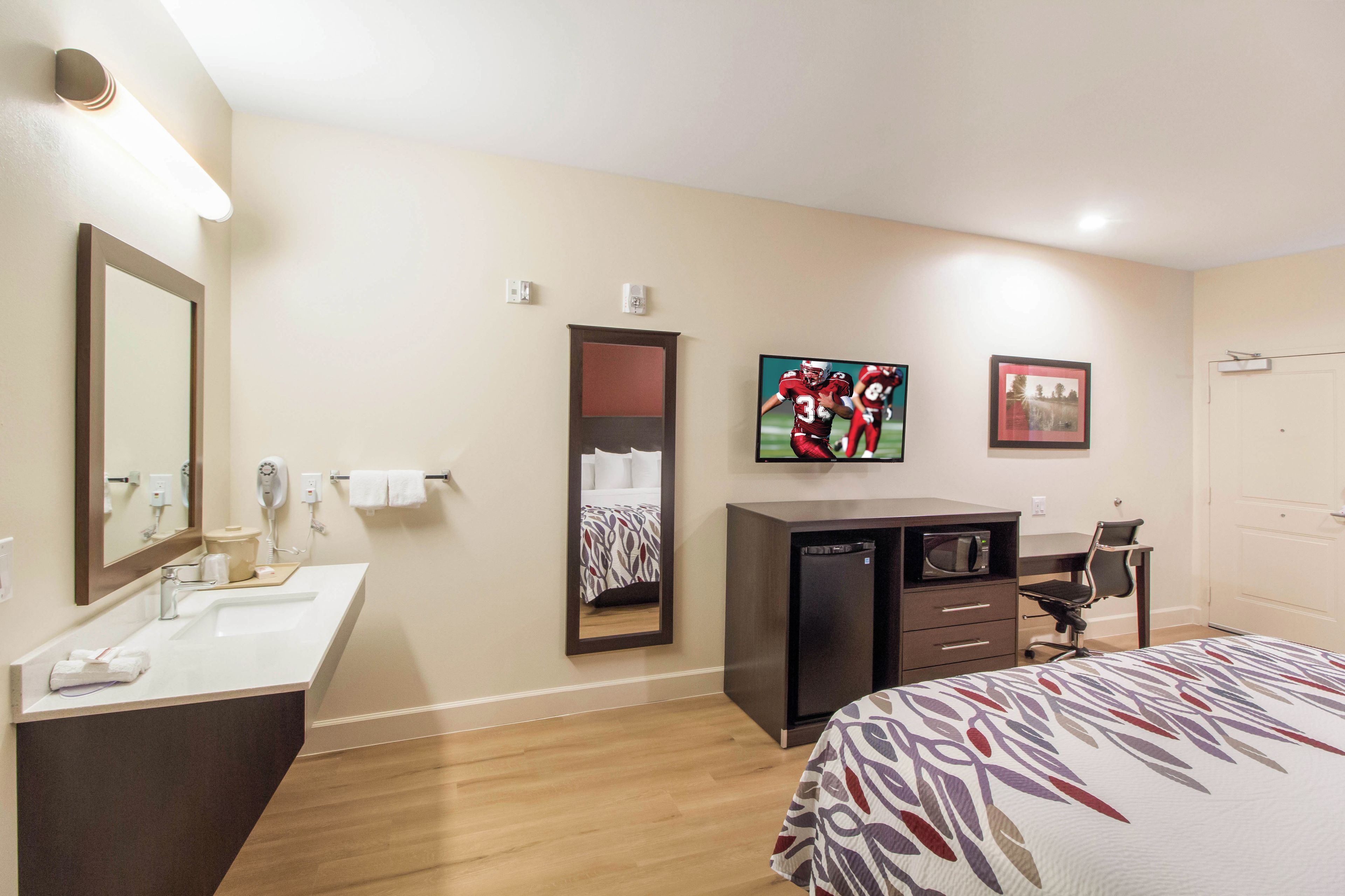 A hotel room with a bed, a desk, a television, a mirror, a bathroom with a sink, and a hairdryer.