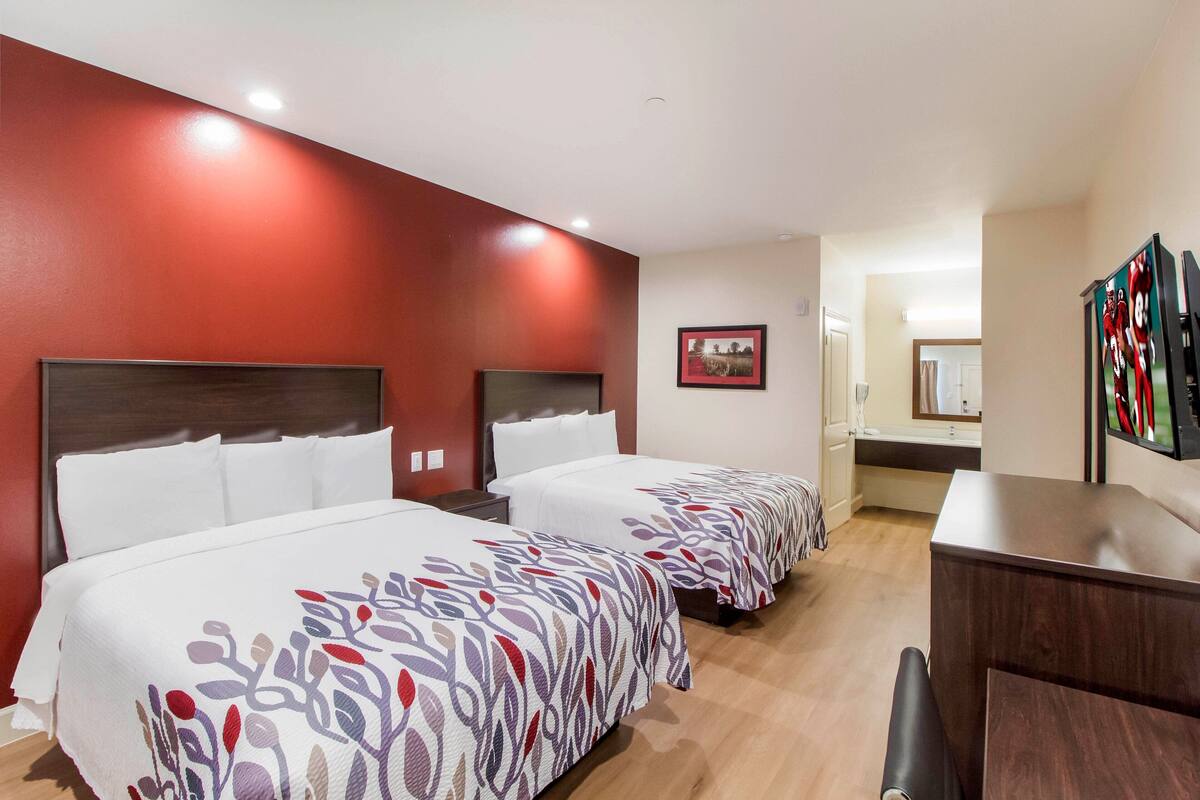 A hotel room with two beds, a desk, and a bathroom visible in the background.
