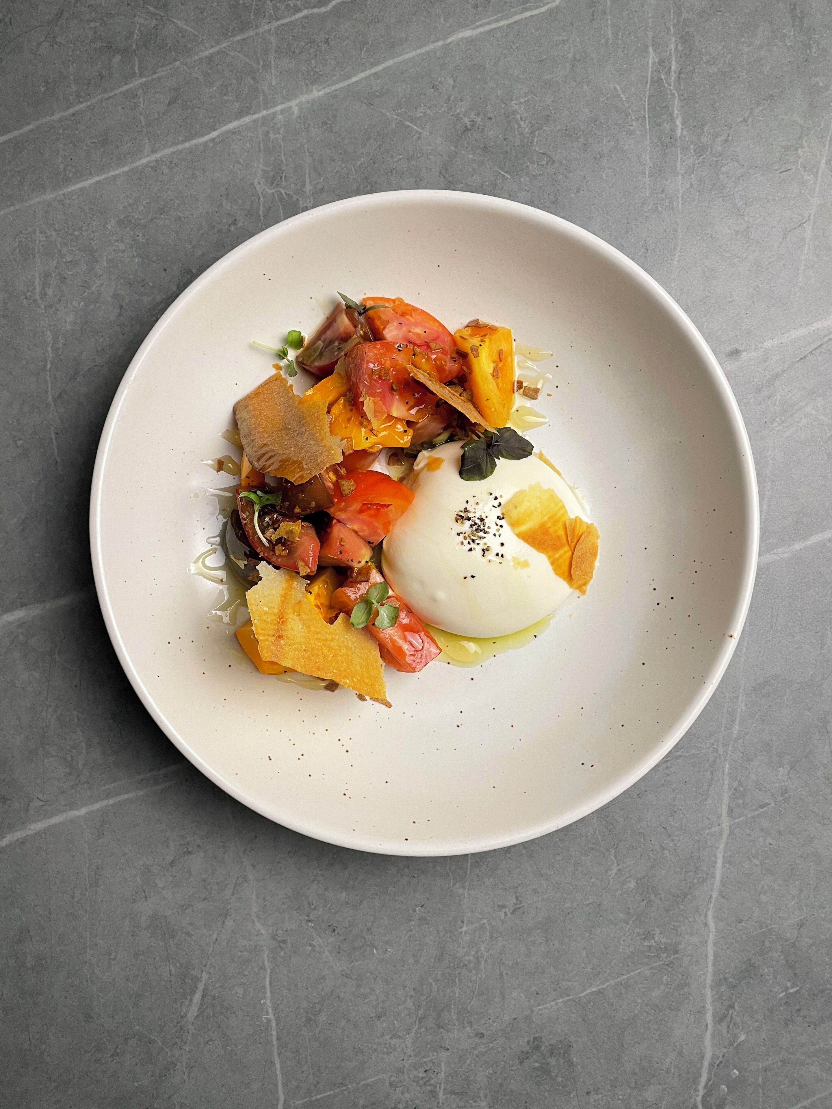 Ein weißer Teller mit pochiertem Ei, gerösteten Tomaten und einem Stück Brot auf einer grauen, strukturierten Oberfläche.
