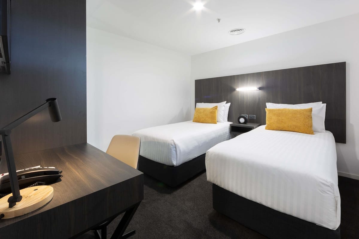 A hotel room with two beds, a desk, a chair, and a lamp.