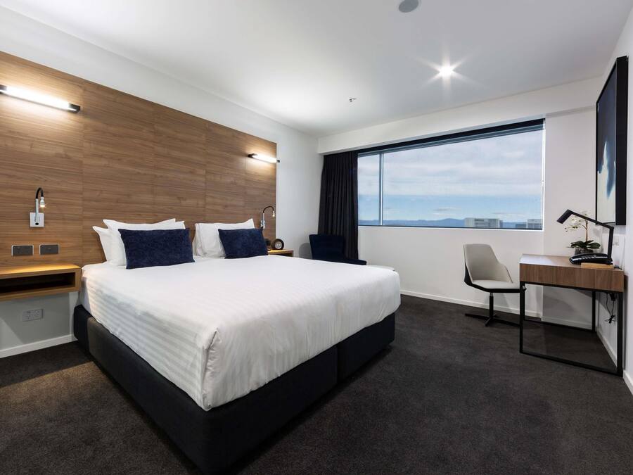 A modern hotel room with a large bed, a desk, a chair, and a view of the city.