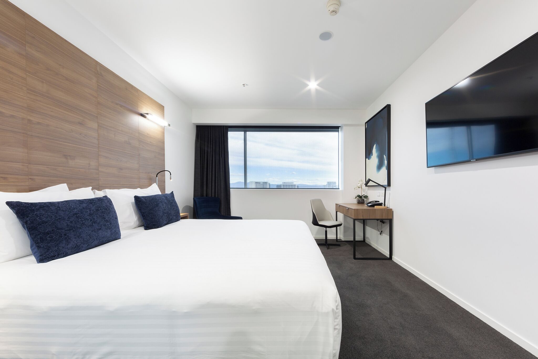A modern hotel room with a large bed, a desk, a chair, a TV, and a window with a city view.