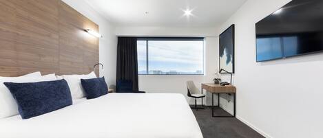 Una habitación de hotel moderna con una cama grande, un escritorio, una silla, un televisor y una ventana con vistas a la ciudad.
