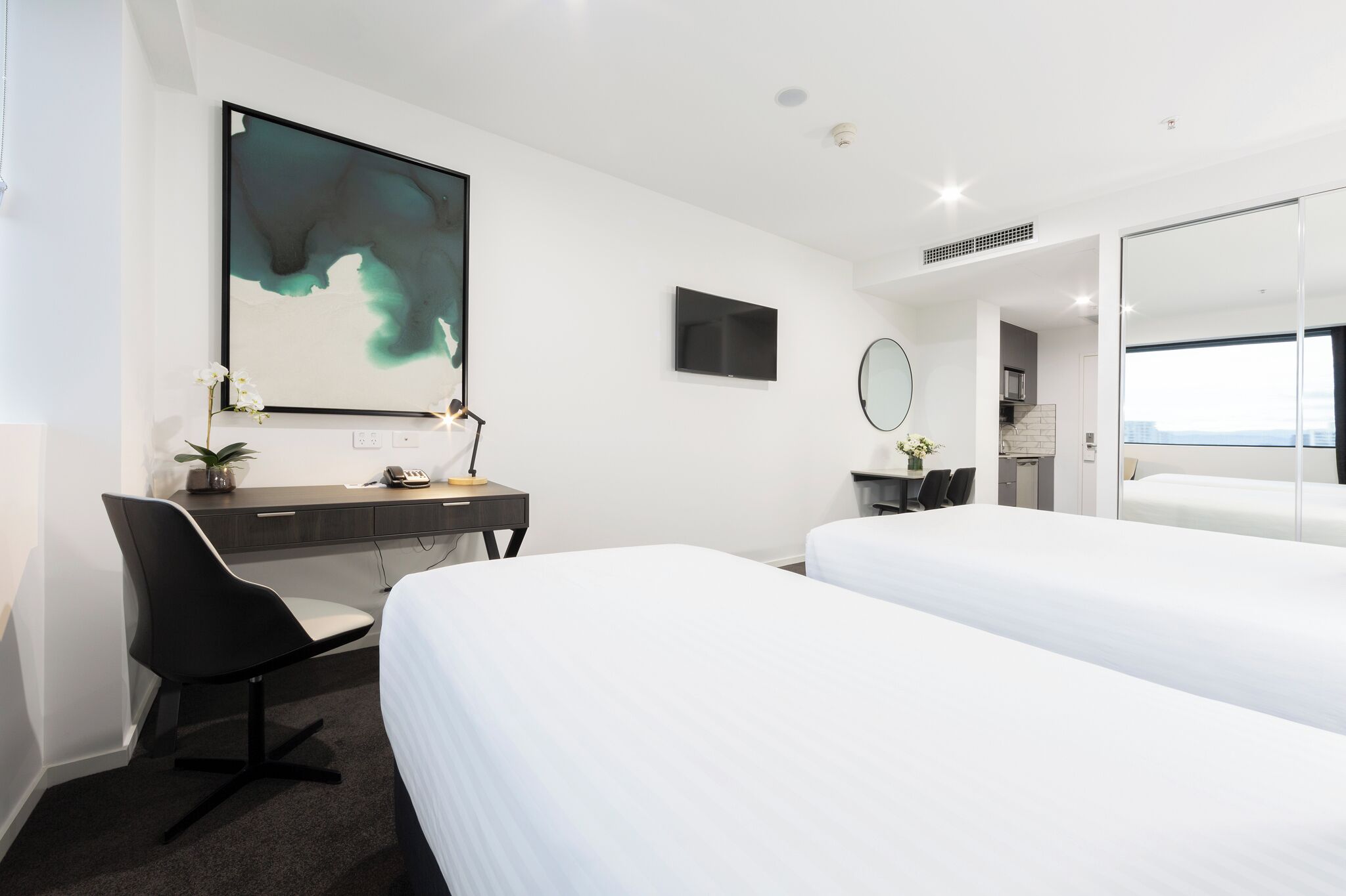 Une chambre d’hôtel moderne avec deux lits, un bureau avec une chaise, une grande peinture abstraite, un téléviseur à écran plat et un grand miroir.