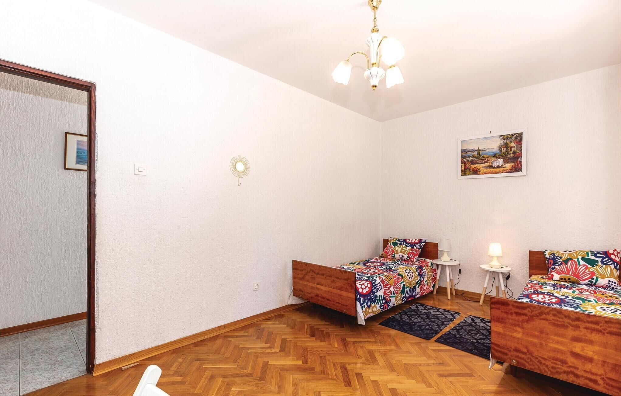 A room with two wooden beds, a small table, a chair, a ceiling light, and a painting on the wall.