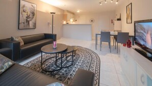Een moderne woonkamer met een zwarte bank, een salontafel en een televisie. Er is een eethoek met een tafel en stoelen. Achtergrond is een keuken.