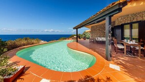 Una piscina con borde curvo, un patio con mobiliario exterior y vista al océano.