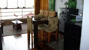 Una habitación con una mesa de comedor, sillas, una mesita con un florero, un sofá, un mueble con una cafetera y una ventana con cortinas.