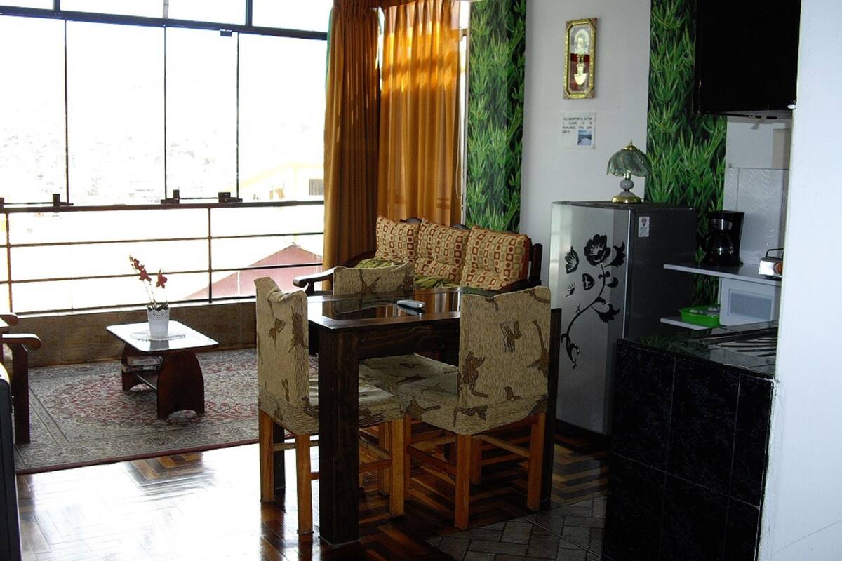 Une pièce comprenant une table à manger, des chaises, une petite table avec un vase, un canapé, un meuble contenant une machine à café et une fenêtre avec des rideaux.