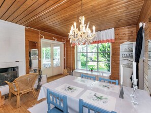 A wooden cabin interior with a dining table, chairs, a chandelier, a fireplace, and a large window with a view of greenery.