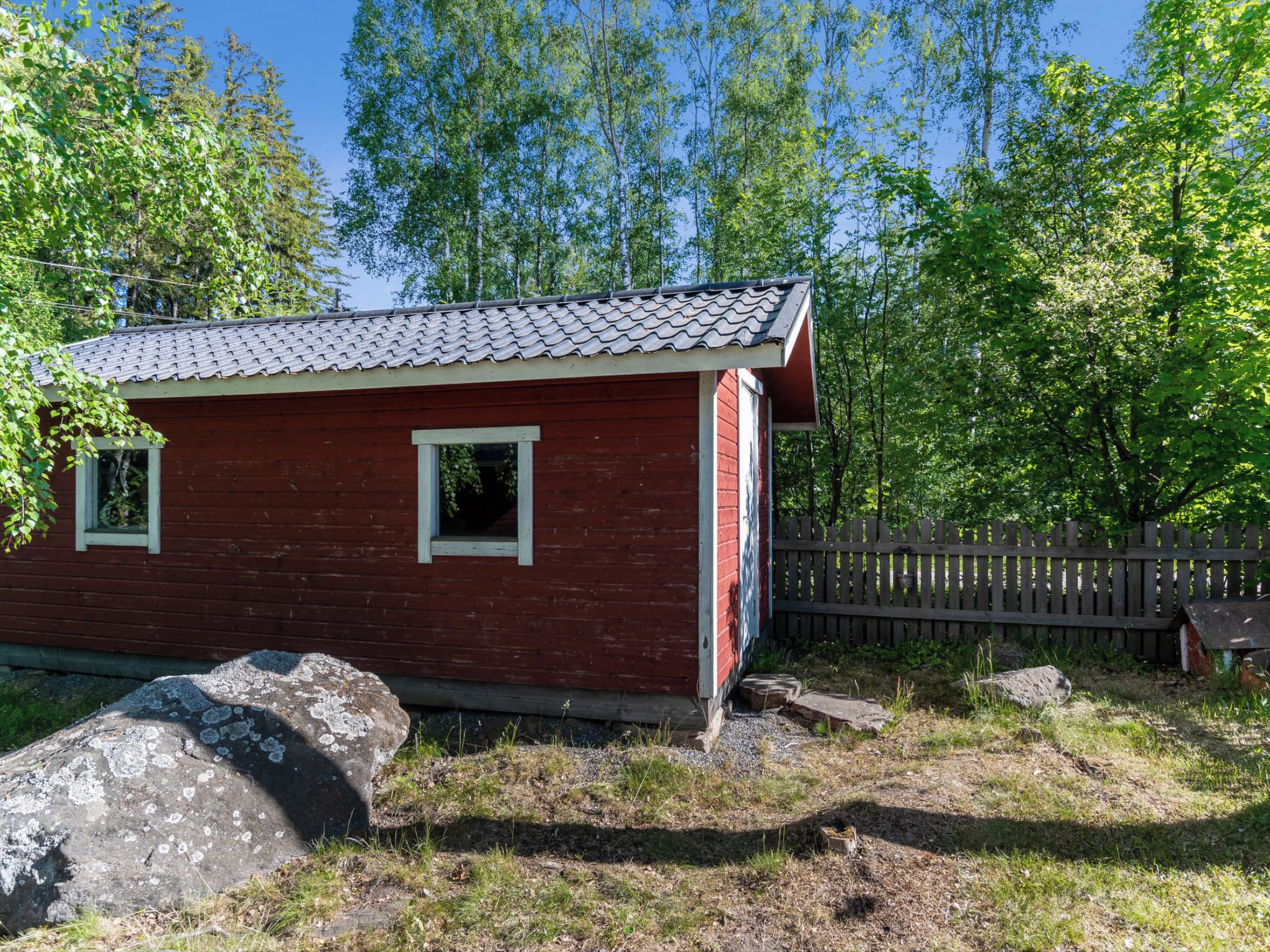 Une petite cabane en bois rouge, avec un toit en tuiles, entourée d’arbres et d’une clôture en bois.