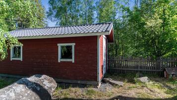 Une petite cabane en bois rouge, avec un toit en tuiles, entourée d’arbres et d’une clôture en bois.