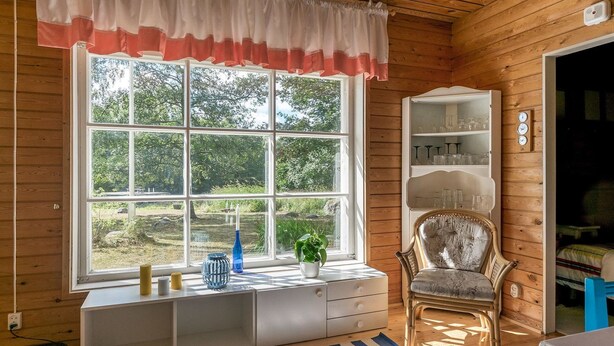 Una habitación de cabaña acogedora con un ventanal grande, un mueble blanco con cajones, una silla y una estantería con cristalería.