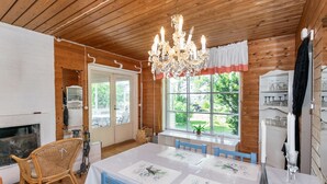 Una cabaña de madera con comedor que incluye una mesa blanca con sillas azules, una araña y un ventanal con vista a follaje.