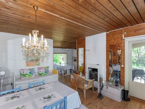 A room with a wooden ceiling, a chandelier, a dining table with chairs, a sofa with cushions, a fireplace, and a window with a view.