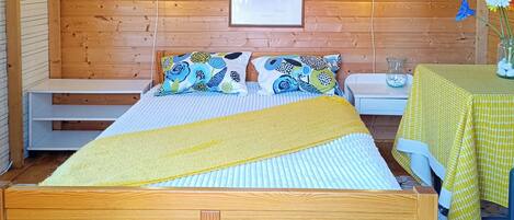 A wooden room with a bed, two bedside tables, a small table with a yellow tablecloth, and a framed picture on the wall.